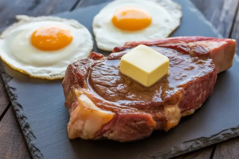 A plate showcasing the 80/20 Carnivore Diet Rule, featuring a fatty ribeye steak with melting butter and two fried eggs, representing a high-fat, moderate-protein meal.